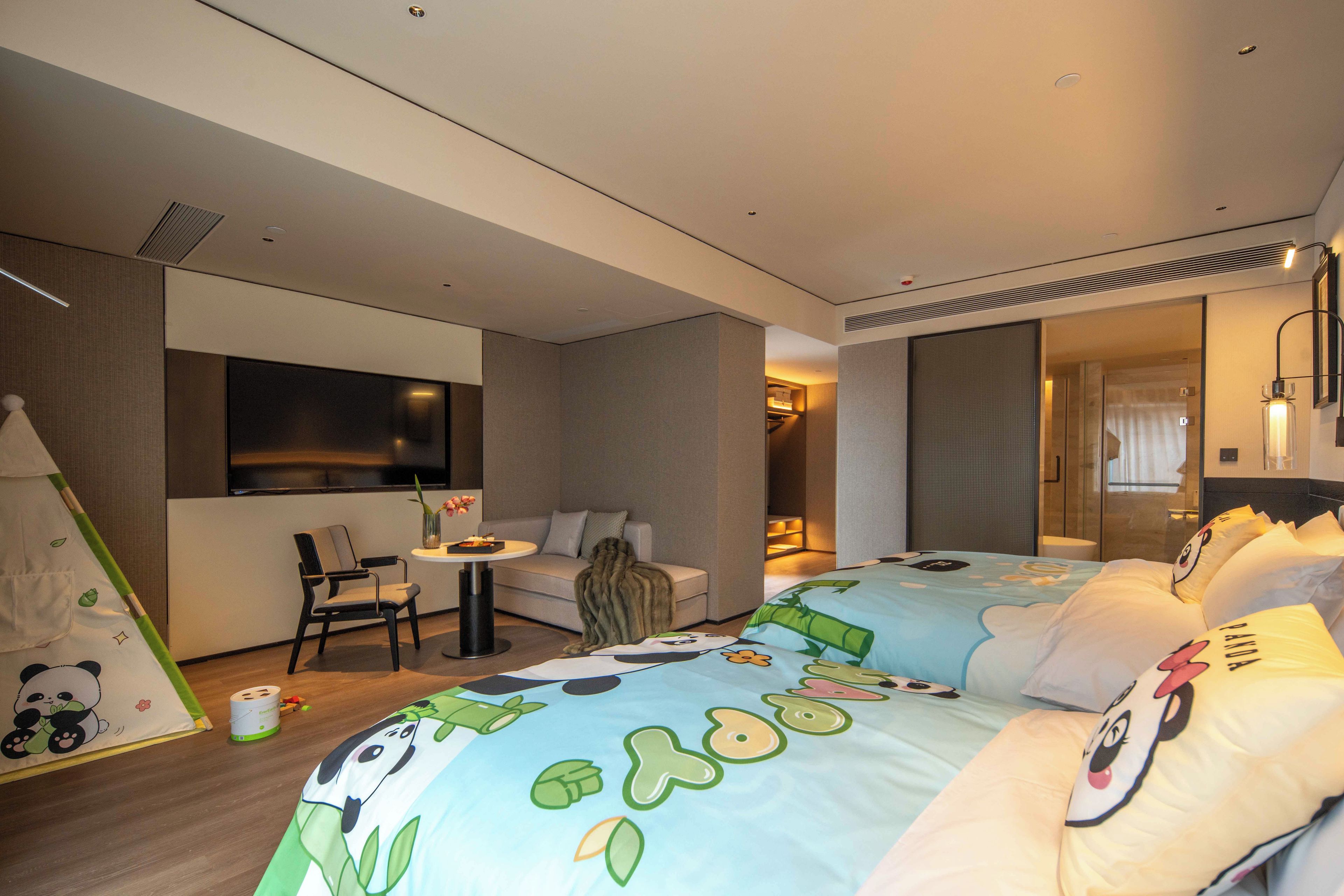 A modern hotel room with a sofa, a small table, a TV, and a bed with a panda-themed bedspread.