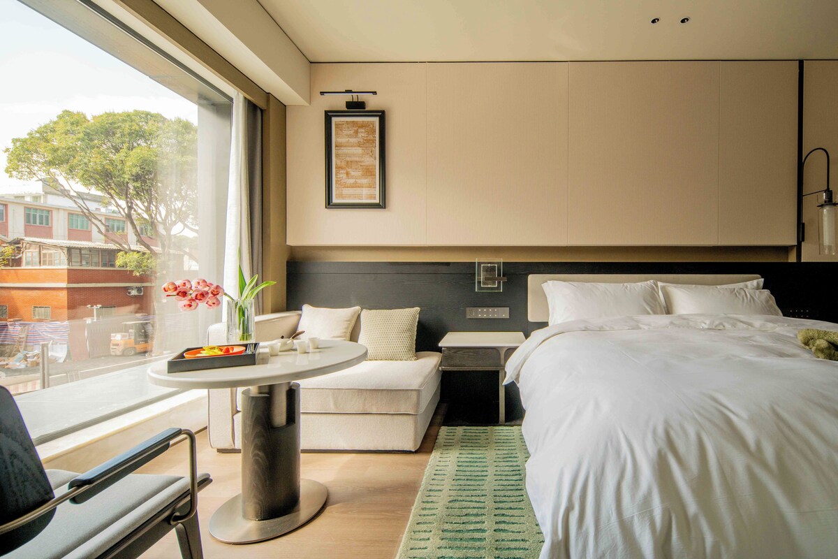 A hotel room with a bed, a small table with a vase of flowers, a chair, a desk, and a window with a view of buildings outside.