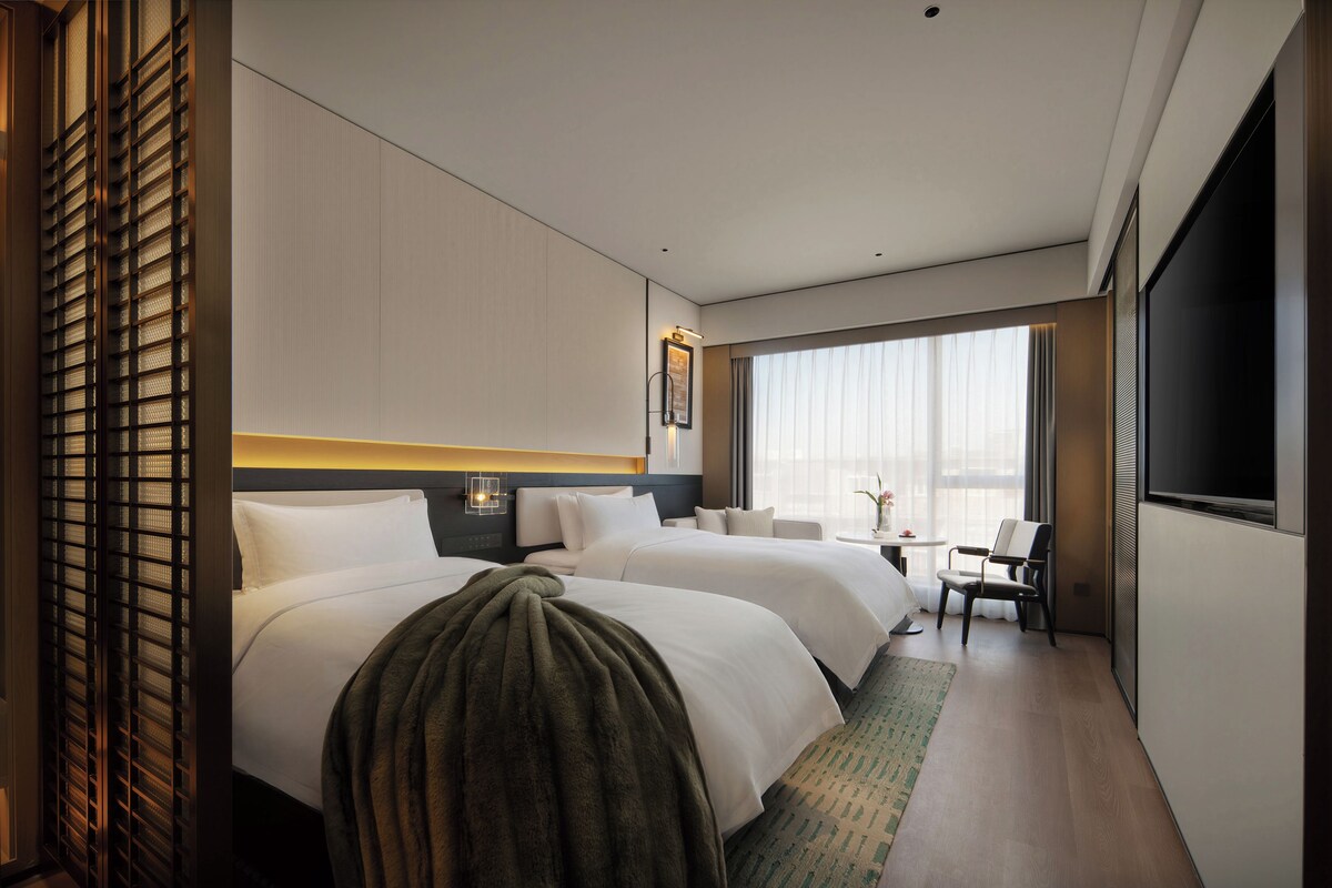 A hotel room with two beds, a sitting area, a television, and a large window.