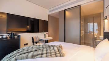 Habitación de hotel moderna con cama, escritorio, silla, televisor y baño visible a través de un vidrio.