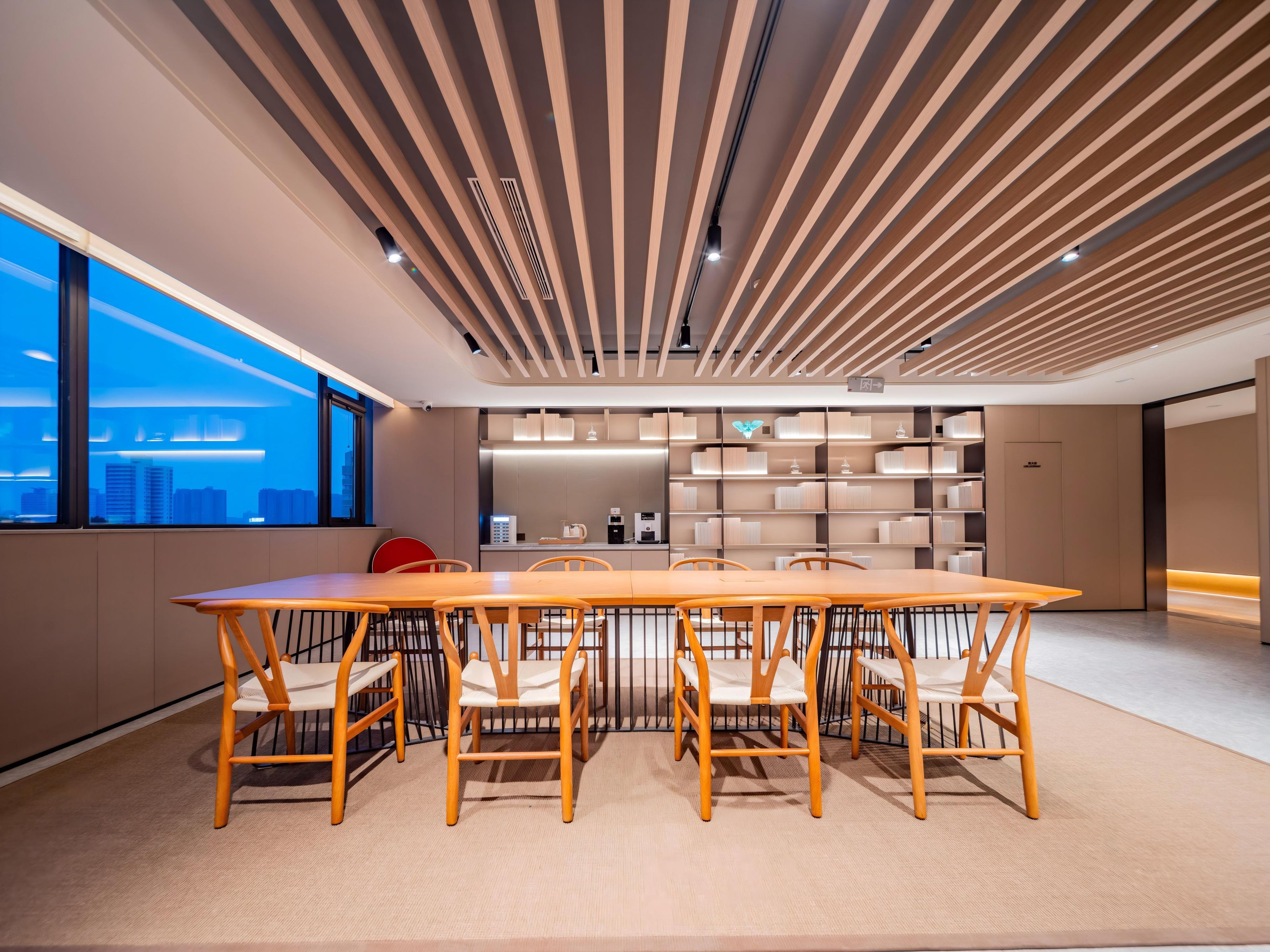 A modern interior with a long wooden table and chairs, a city view through large windows, and a ceiling with wooden slats.