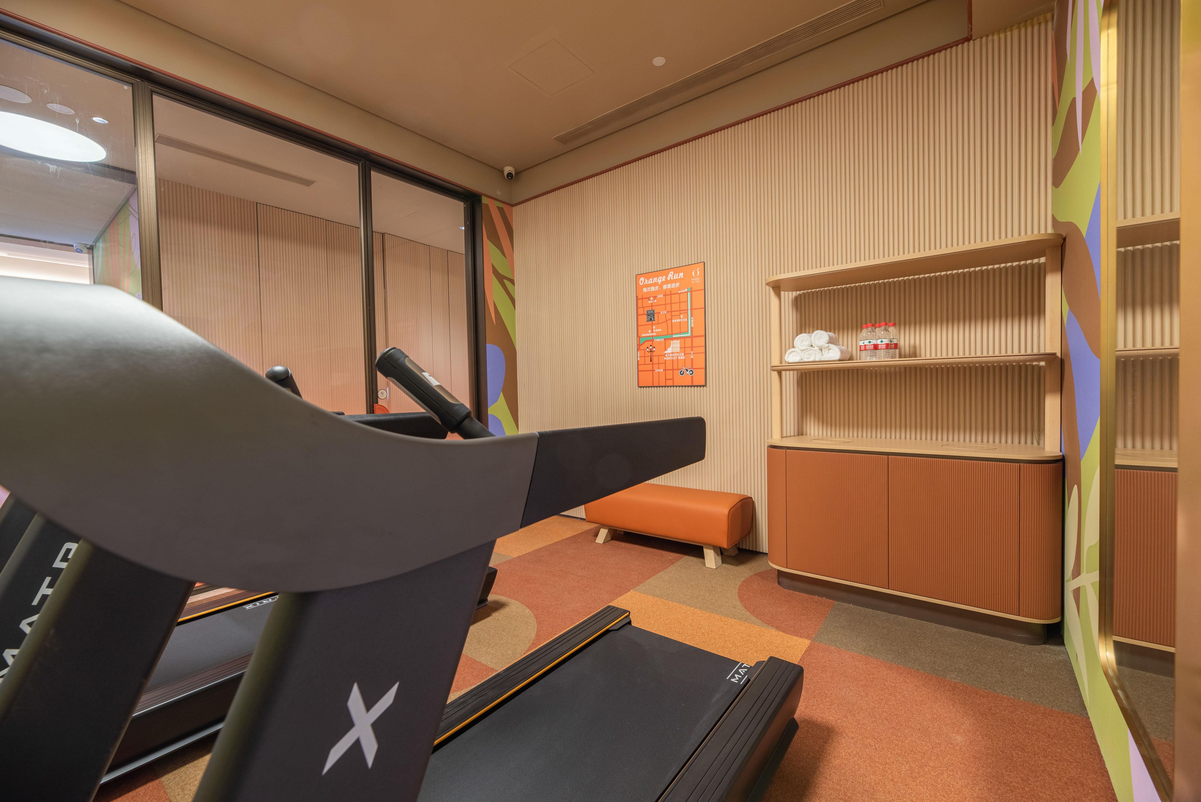 A hotel room with a treadmill, a small orange bench, a wall-mounted shelf with toiletries, and a map on the wall.