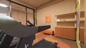 A hotel room with a treadmill, a small orange bench, a wall-mounted shelf with toiletries, and a map on the wall.