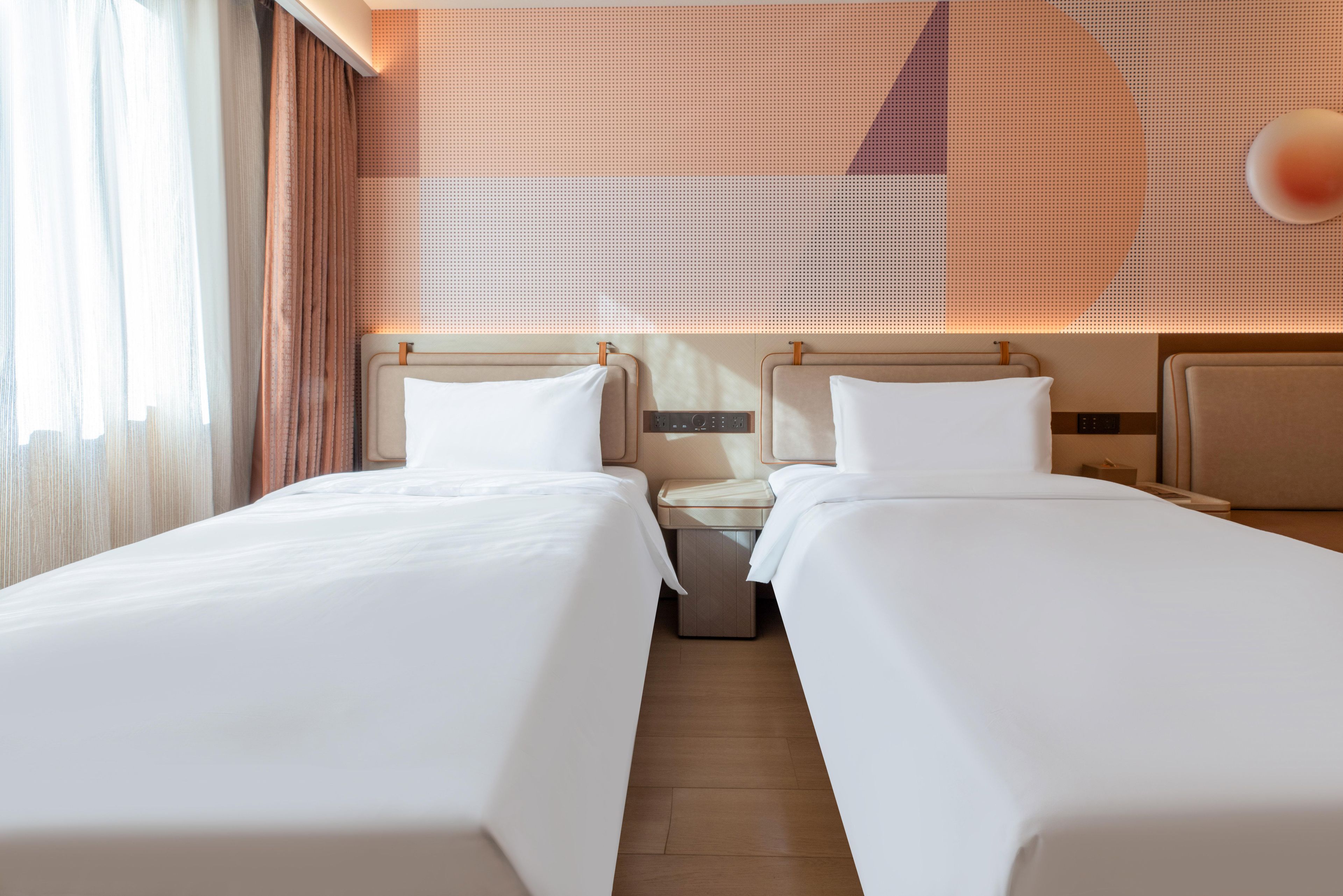 A hotel room with two single beds, a nightstand, a wall-mounted control panel, and a large window with curtains.