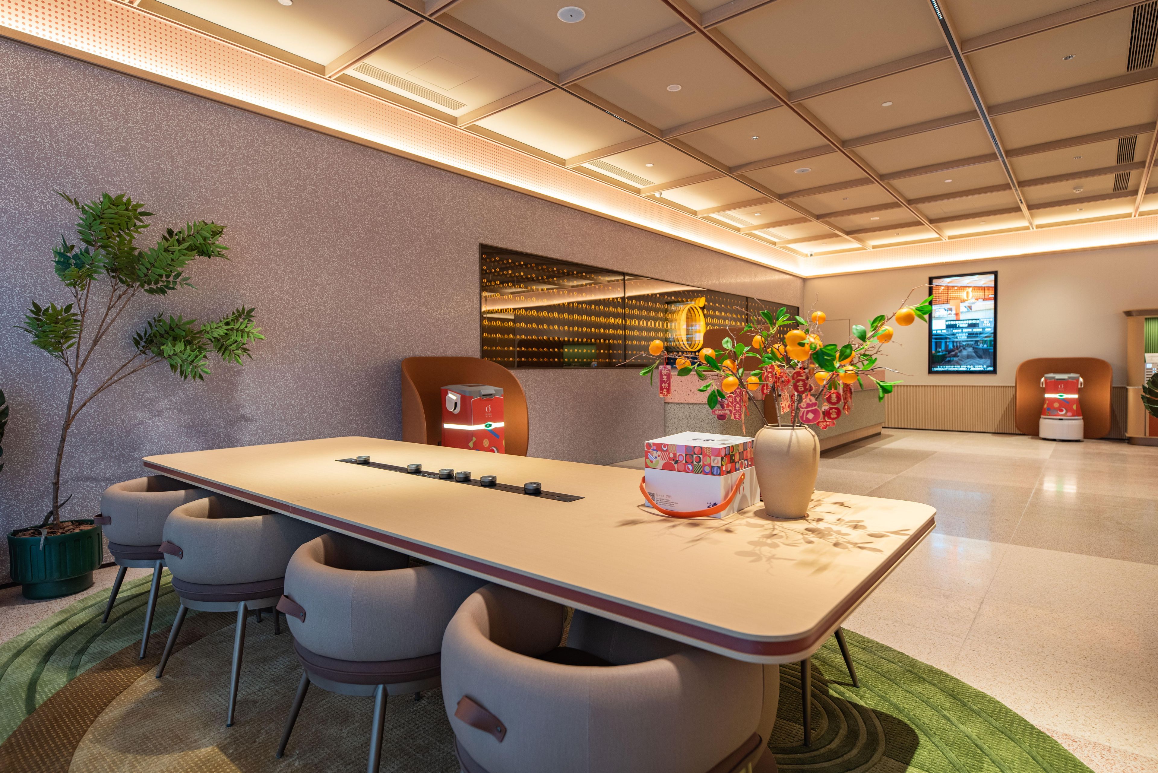 A modern hotel lobby with a long reception desk, comfortable seating, a large vase with flowers, and a digital screen displaying information.