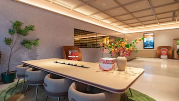 A modern hotel lobby with a long reception desk, comfortable seating, a large vase with flowers, and a digital screen displaying information.