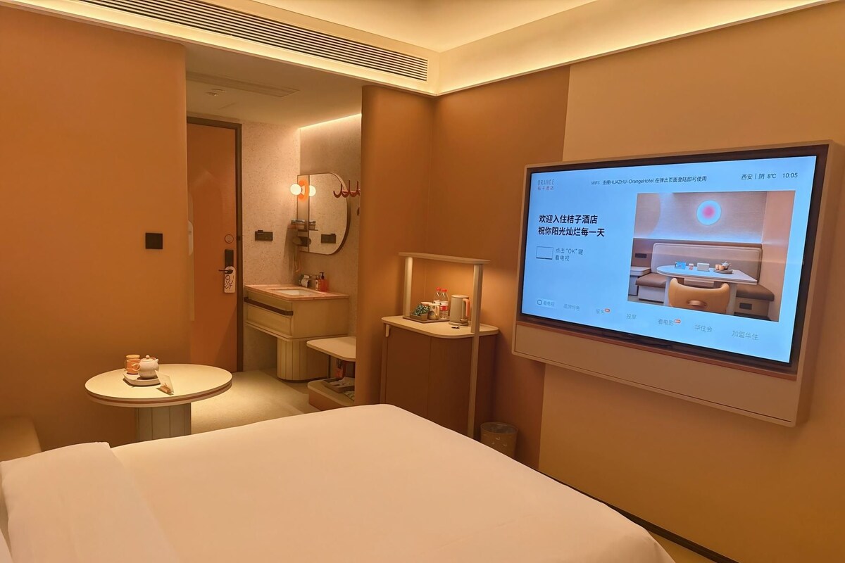 Ein Hotelzimmer mit Bett, einem kleinen Tisch mit Teekanne, einem Spiegel und einem an der Wand befestigten Fernseher, der einen Hotelinnenraum zeigt.