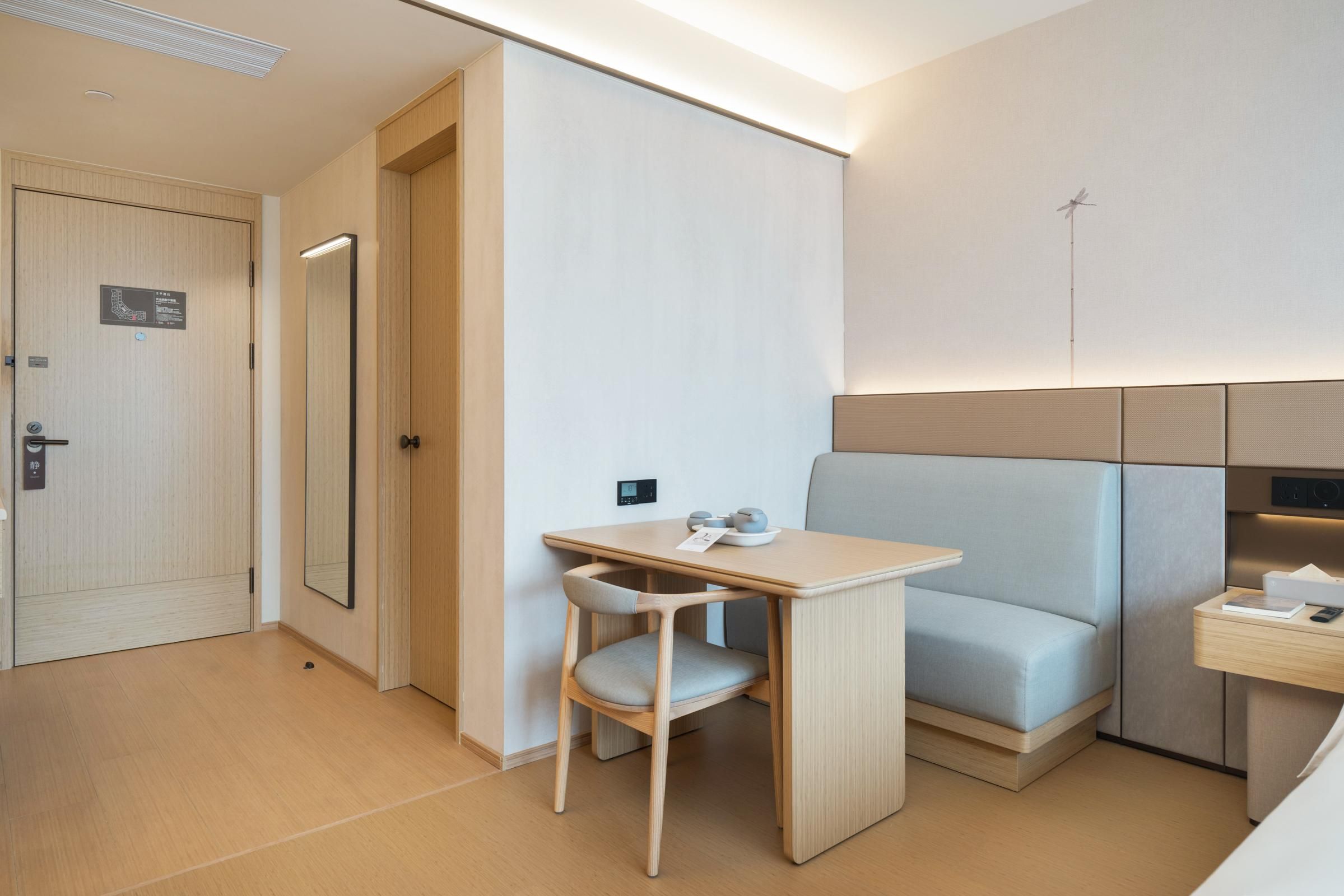 Una habitación de hotel moderna con una puerta de madera, una mesita con una silla, una cama con cabecero y una mesita de noche con lámpara.