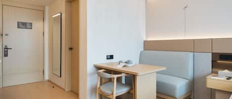 Quarto de hotel moderno com porta de madeira, uma pequena mesa com cadeira, cama com cabeceira e mesa de cabeceira com abajur.