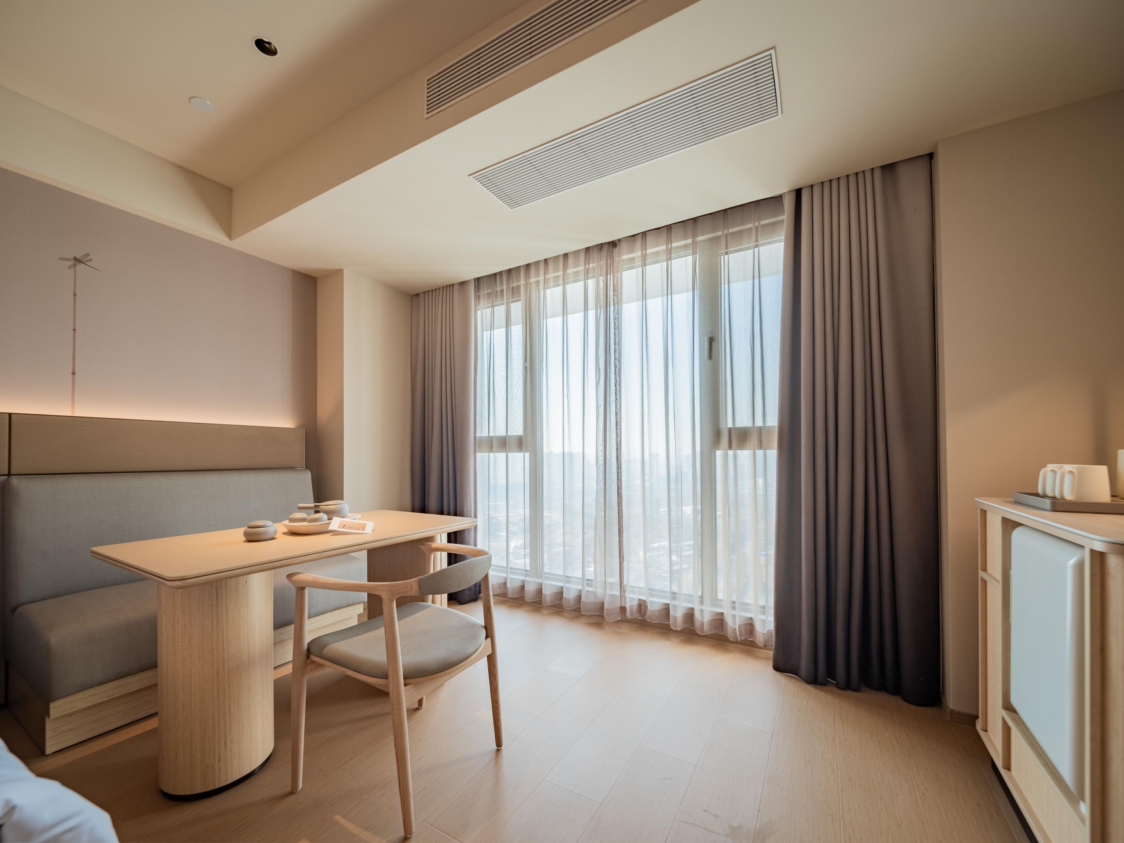 A modern hotel room with a wooden desk, a cushioned bench, a chair, and a cabinet.