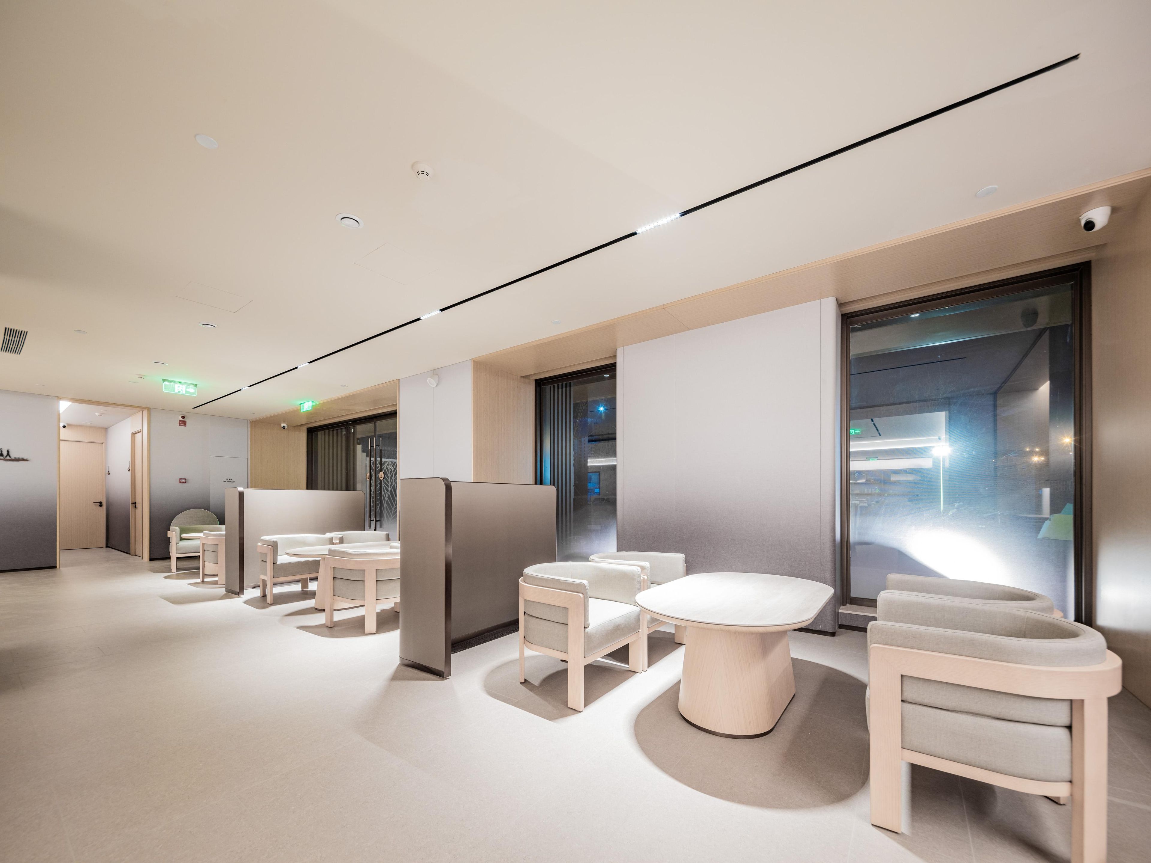 A modern hotel lobby with a minimalist design, featuring a central seating area with round tables and chairs, a glass partition wall, and a ceiling with recessed lighting.