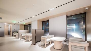 A modern hotel lobby with a minimalist design, featuring a central seating area with round tables and chairs, a glass partition wall, and a ceiling with recessed lighting.