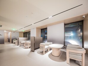 A modern hotel lobby with a minimalist design, featuring a central seating area with round tables and chairs, a glass partition wall, and a ceiling with recessed lighting.