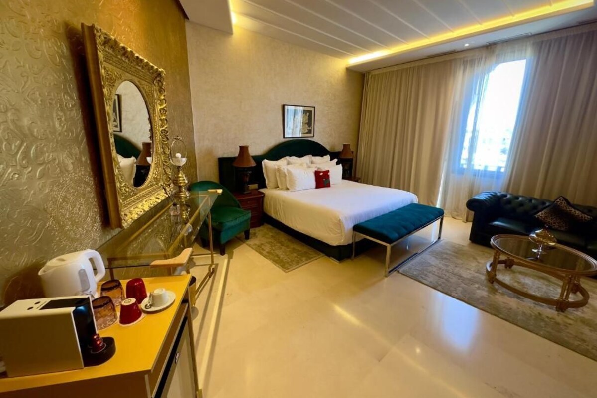 Habitación de hotel con cama, escritorio con cafetera, espejo, sofá y ventana grande con cortinas.