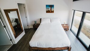 A bedroom with a large bed, wooden flooring, a door with a mirror, a bedside table with a lamp, and a window with a view. - The Spot (Winnipeg)