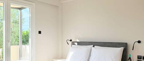 A hotel room with a bed, a bedside table with a bottle and glasses, and a view of greenery outside.