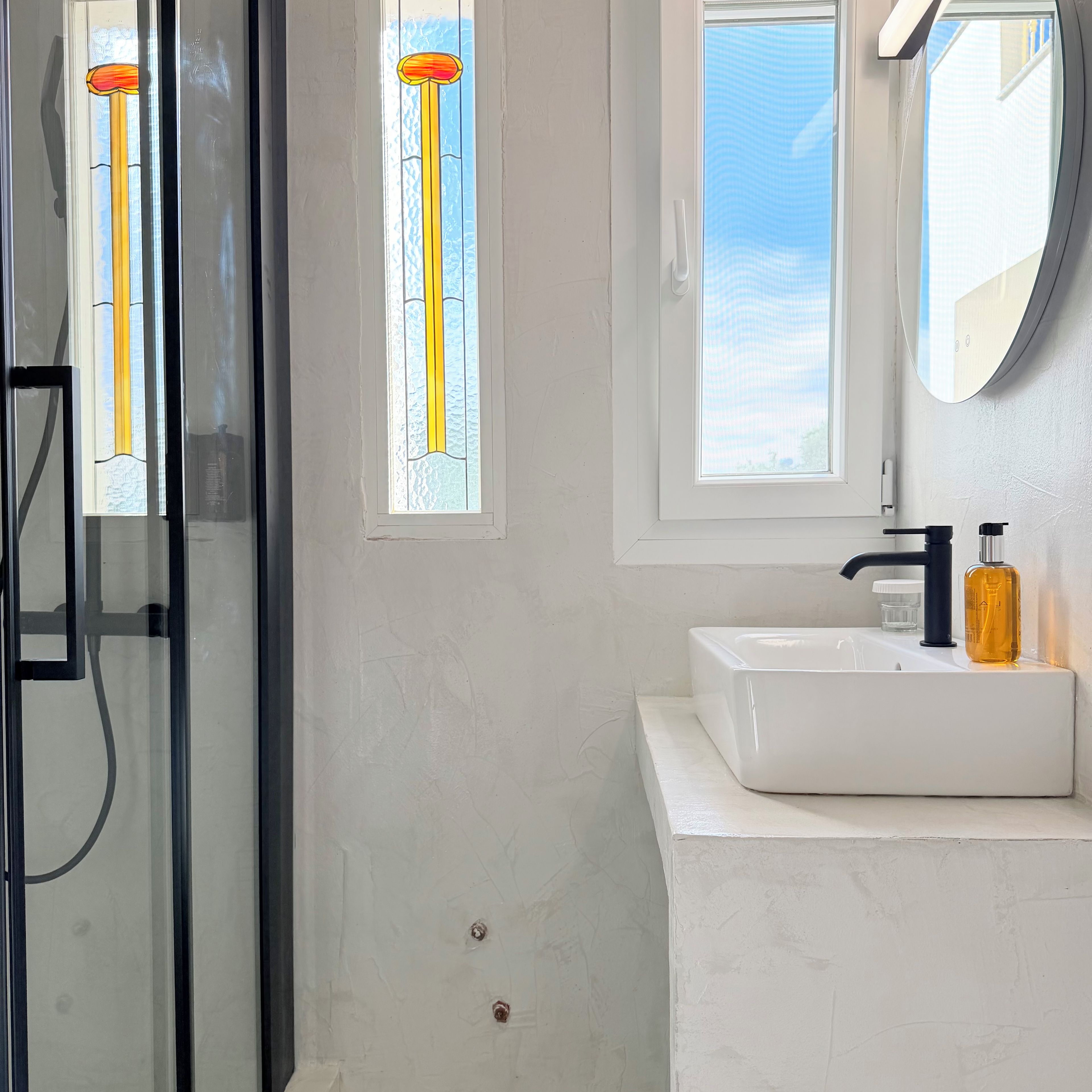 A bathroom with a glass shower enclosure, a white sink with a black faucet, and a window with a view of the sky.