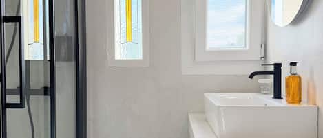 A bathroom with a glass shower enclosure, a white sink with a black faucet, and a window with a view of the sky.
