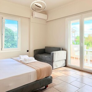 A bedroom with a bed, a grey sofa, a large window, and an air conditioning unit. - Altamar Collection (Istiaia-Aidipsos)