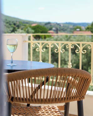 A wicker chair and a table with a glass of white wine on a balcony with a metal railing and a view of greenery and buildings.