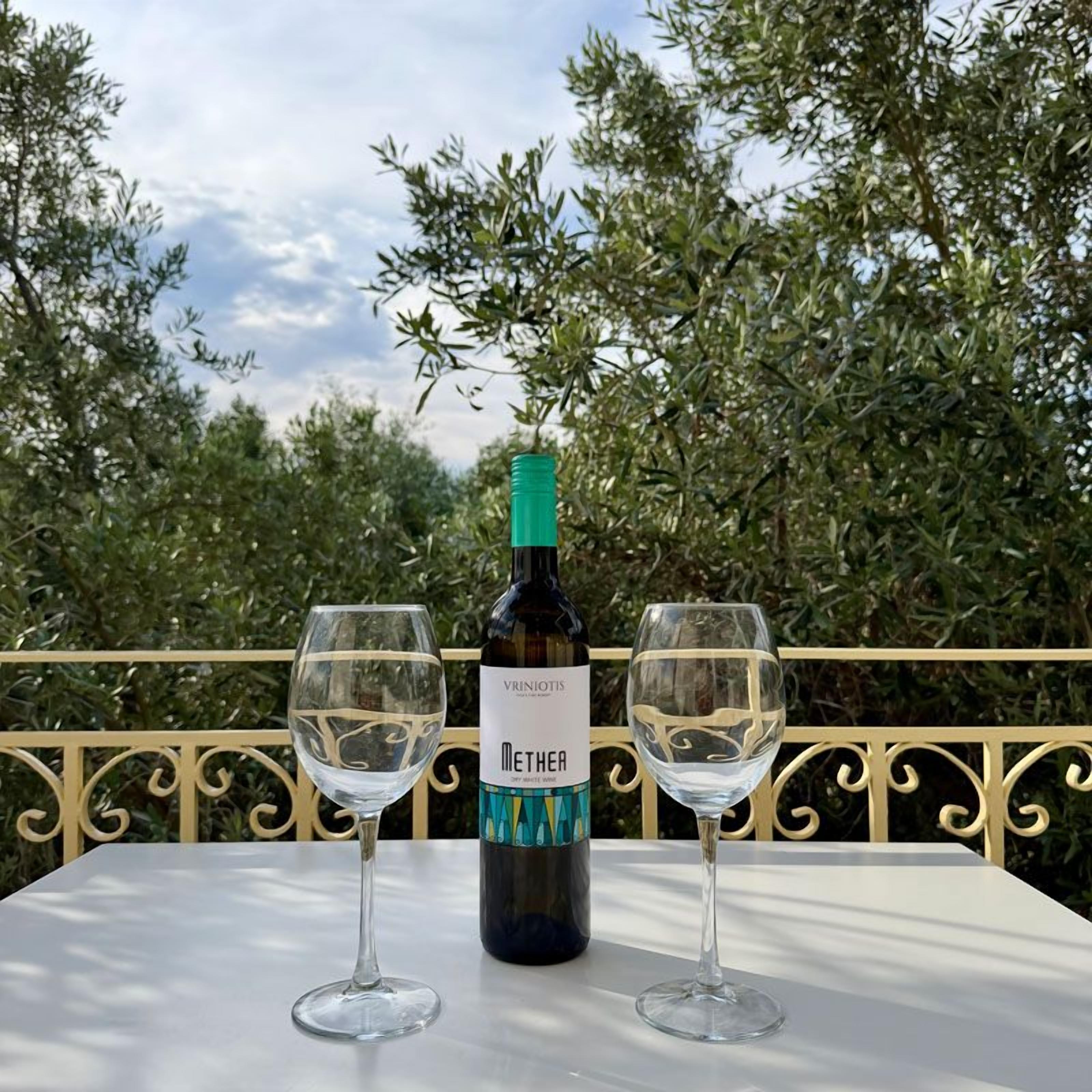 Una botella de vino Vrinchiotis Metha y dos copas de vino vacías sobre una mesa con vistas a olivares.