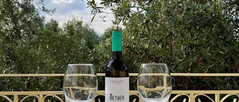 Una botella de vino Vrinchiotis Metha y dos copas de vino vacías sobre una mesa con vistas a olivares.