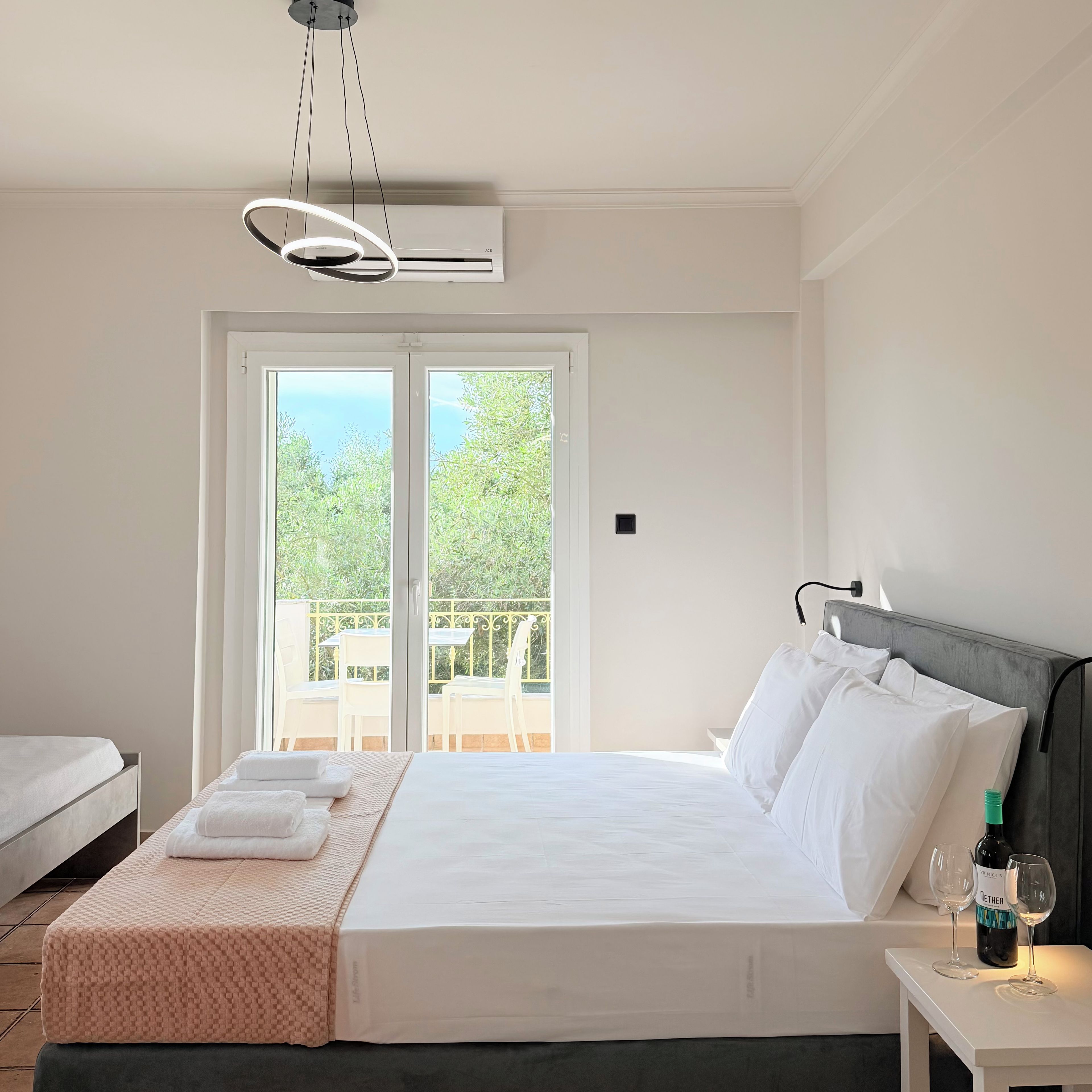 A hotel room with a bed, a bedside table with a bottle and glasses, a window with a view of greenery, and an air conditioning unit.