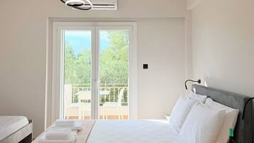 A hotel room with a bed, a bedside table with a bottle and glasses, a window with a view of greenery, and an air conditioning unit.