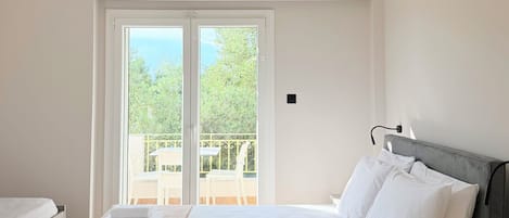 Habitación de hotel con cama, mesita de noche con una botella y vasos, ventana con vistas a zonas verdes y un equipo de aire acondicionado.