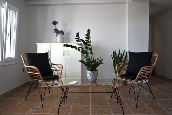 A room with two wicker chairs, a glass table, a cabinet, and potted plants.
