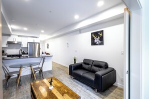 A modern kitchen with stainless steel appliances, a dining area with a glass table and metal chairs, a black leather sofa, and a deer artwork on the wall. - Blvd Beltline (Calgary)