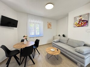 Una sala de estar moderna con un sofá gris, una mesa de centro redonda de madera, una mesa de comedor con sillas, un televisor de pantalla plana montado en la pared y una ventana con cortinas traslúcidas.