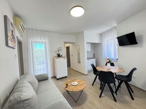 Una sala de estar moderna con un sofá, una mesa de comedor con sillas, una zona de cocina y un televisor montado en la pared.