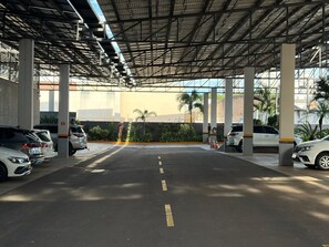 Area di parcheggio coperta con diverse auto parcheggiate, pilastri in cemento e cielo sereno.