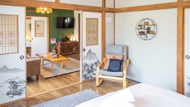 Une pièce de style japonais traditionnel, avec un sol recouvert de tatamis, un lit bas en bois, un fauteuil avec des coussins, une télévision et une petite table avec une lampe.