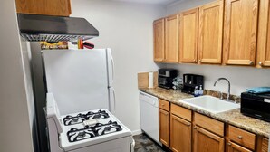 Una cocina compacta con estufa a gas, lavavajillas, armarios de madera y encimera de granito.