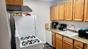 Una cocina compacta con estufa a gas, lavavajillas, armarios de madera y encimera de granito.