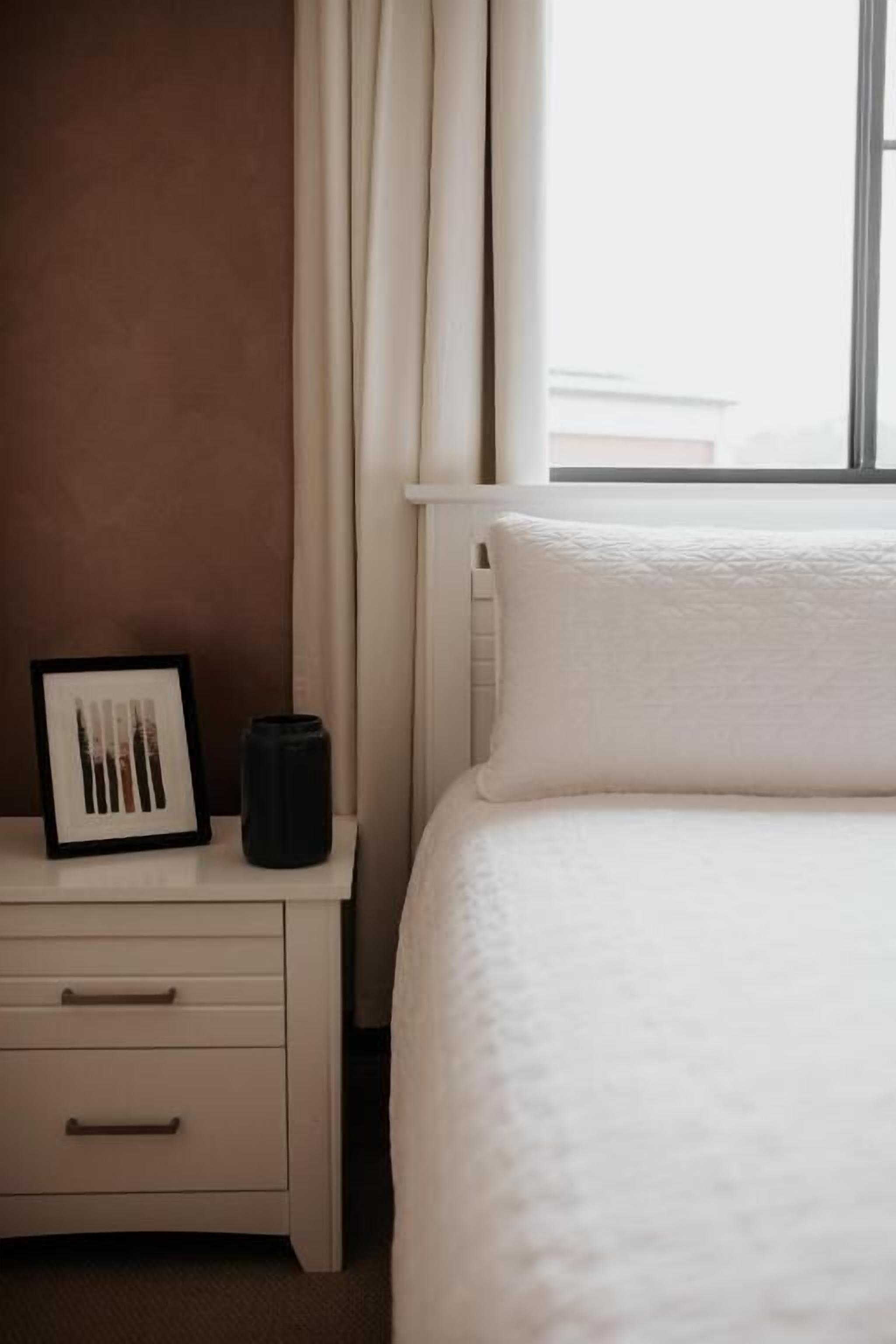 Una cama bien hecha con una colcha blanca, una mesita de noche con un cuadro enmarcado y un jarrón negro, y una ventana con cortinas.