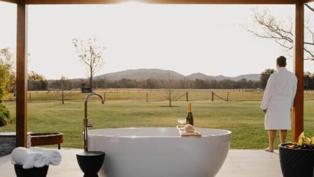 A modern outdoor bathroom with a large bathtub, a towel rack, and a potted plant.