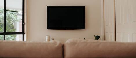 Un saló amb un sofà beix, una televisió plana penjada a la paret, una planta petita i una finestra amb vistes a la vegetació.