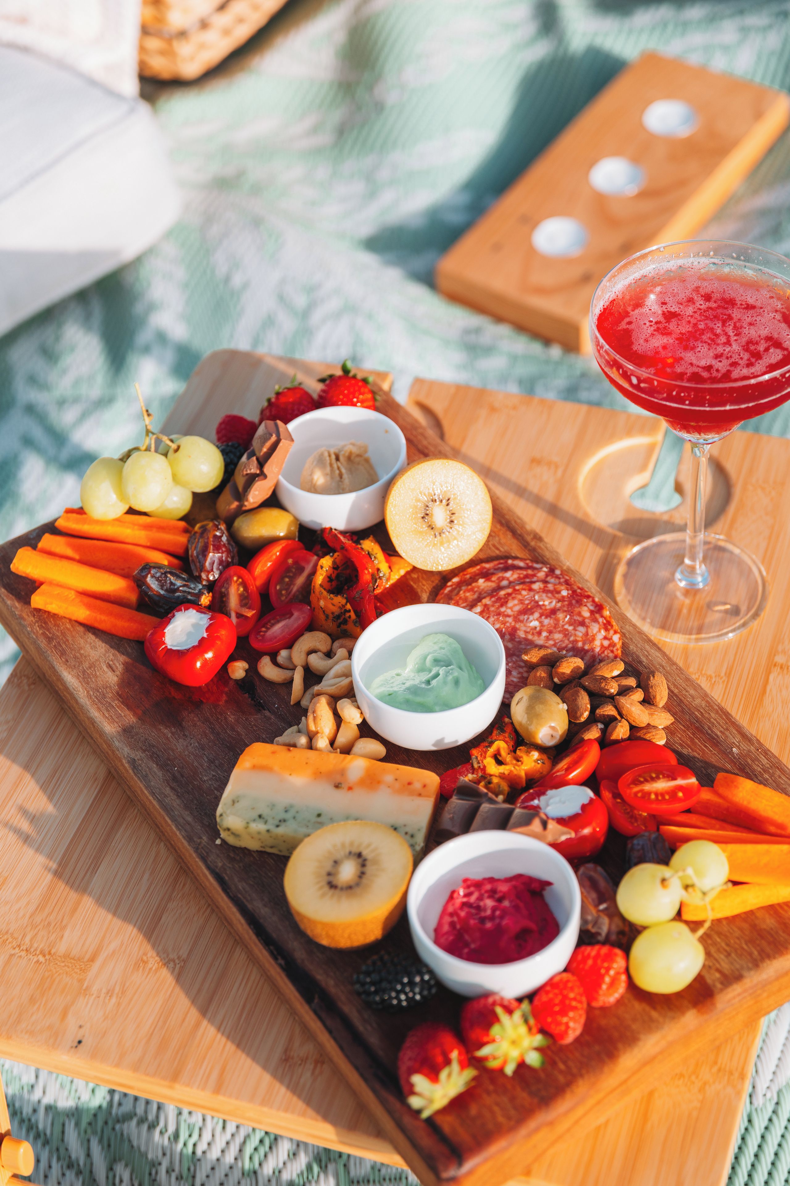 A wooden board with a variety of cheeses, fruits, nuts, and dips, accompanied by a glass of red drink.