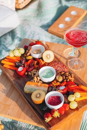 Una tabla de madera con varios quesos, frutas, frutos secos y salsas, acompañada de una copa de bebida roja.