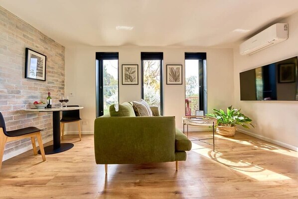 Un salon moderne avec un canapé vert, une table à manger avec des chaises, une plante en pot et des œuvres d’art encadrées accrochées aux murs.