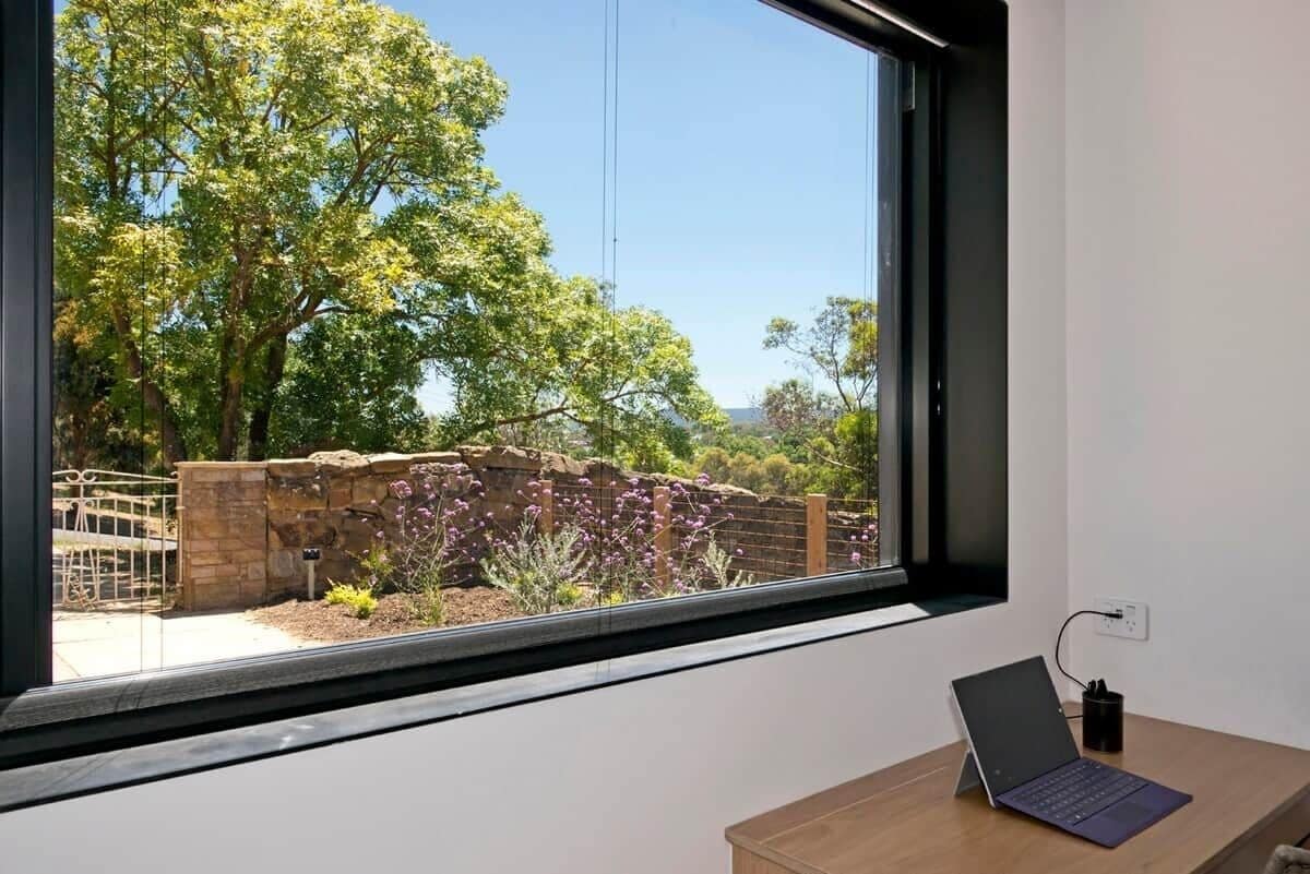 A laptop on a desk near a window overlooking a garden with trees and a stone wall.
