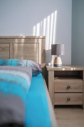 Ein Schlafzimmer mit einem hölzernen Kopfteil, einem Bett mit blauer und grauer Bettdecke, einem Nachttisch mit Lampe sowie einem Schubladenschrank.