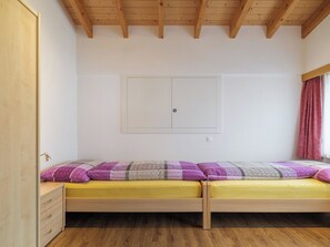Ein Zimmer mit Holzboden, einem Bett mit gelber Matratze und violetter Bettwäsche, einem Holzschrank und einem Fenster mit rotem Vorhang.