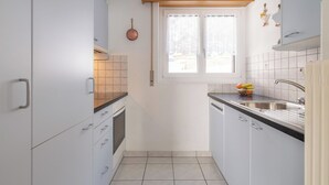 Et lille køkken med hvide skabe, indbygget ovn, et vindue med gardiner og en vask med hane.