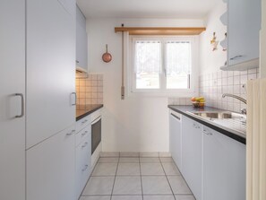 A compact kitchen with white cabinets, a built-in oven, a window with curtains, and a sink with a faucet. - Alpen Leukerbad # 4-bed apartment, Dusche, WC by Interhome (Leukerbad)