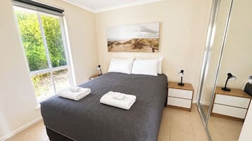 A bedroom with a large bed, a bedside table, a nightstand, a window with a view of greenery, and a painting on the wall.