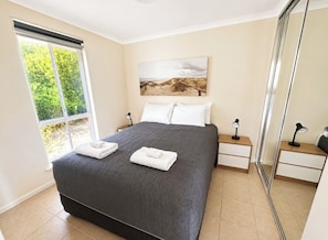 A bedroom with a large bed, a bedside table, a nightstand, a window with a view of greenery, and a painting on the wall.